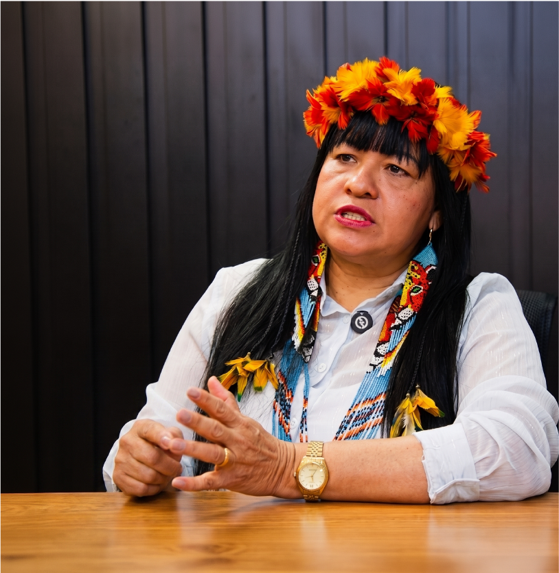 Luciene Kayabi conversando à mesa, com coroa de penas coloridas